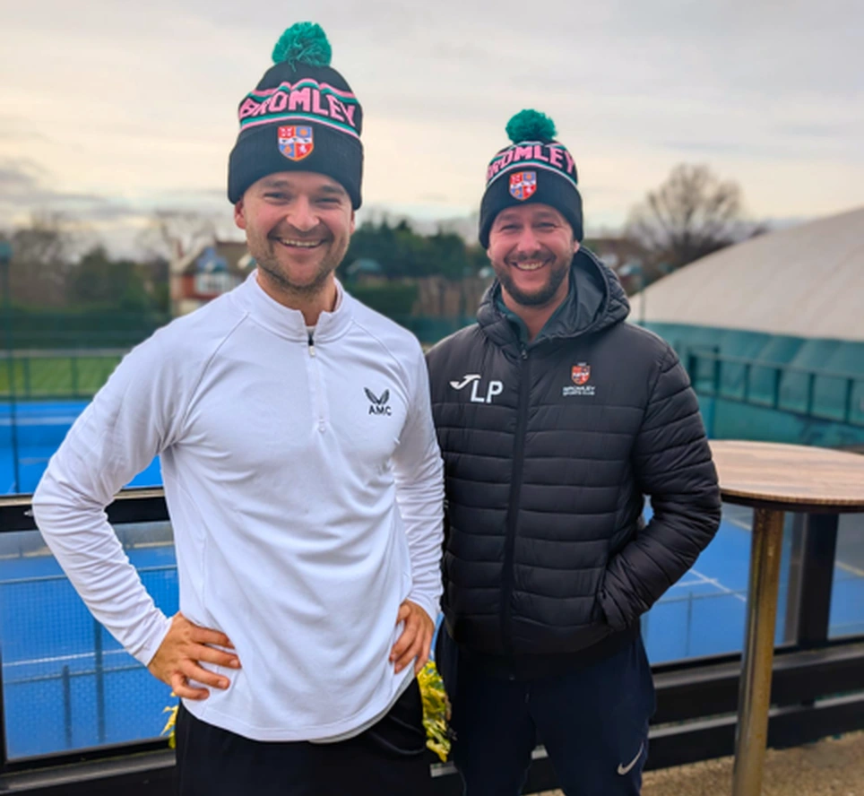 Two Bromley Sports Club coaches wearing official Bromley beanie hats on the tennis courts at the club