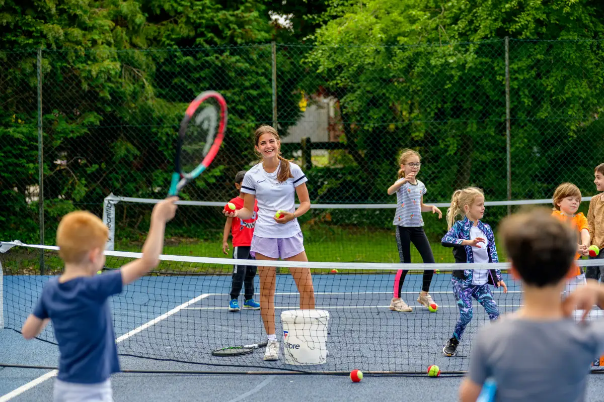 Coach leading a group of young children in a fun tennis session on outdoor courts.